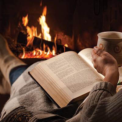 Person with a book on their lap, sitting by the fire, holding a mug